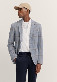 Hellblaues Karo-Blazer mit braunen und marineblauen Akzenten, kombiniert mit einem weißen Hemd und marineblauen Hosen. Das Modell trägt eine beige Mütze.