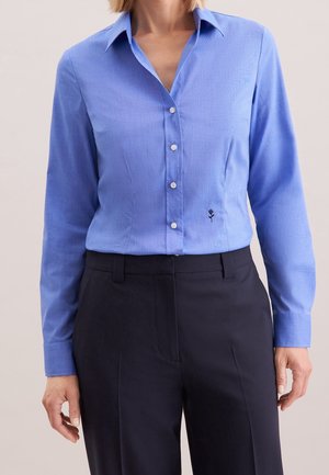 Vrouw die een aansluitend lichtblauw overhemd met lange mouwen en kleine zwarte bloemenborduursels draagt, boven zwarte op maat gemaakte broek.