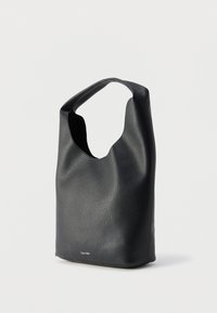 Μαύρη τσάντα ώμου από δερματίνη με φαρδιά λουρί, στρογγυλεμένη κορυφή και μικρό λογότυπο "Calvin Klein" κοντά στο κέντρο του κάτω μέρους.