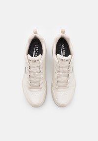 Pár béžových tenisek Skechers se šněrováním, černými polstrovanými stélkami a viditelným názvem značky na jazycích a bocích, pohled shora.