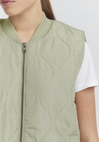 Gestepptes olivgr�ünes ärmelloses Jacket mit Reißverschluss, das dezente wellenförmige Nähte und einen gerippten Kragen aufweist. Darunter ist ein weißes Hemd sichtbar.
