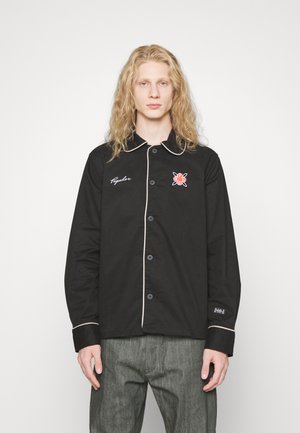 Svart skjorte med knapper, kontrasterende lyst kantbånd, med brodert design på venstre bryst og avslappet passform.