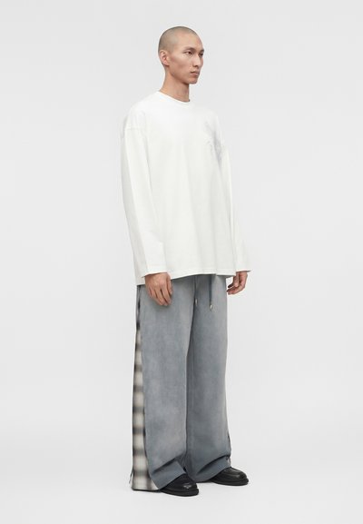 Άνδρας μοντέλο που στέκεται, φοράει υπερμεγέθη άσπρο πουκάμισο μακρυμάνικο και φαρδιά γκρι παντελόνια με ασπρόμαυρα καρό πλαϊνά πάνελ, μαύρα παπούτσια.