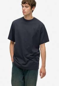 Jonge man met kort bruin haar, gekleed in een losse donkerblauwe T-shirt en blauwe jeans, staande met één hand in zijn zak, kijkend naar links.