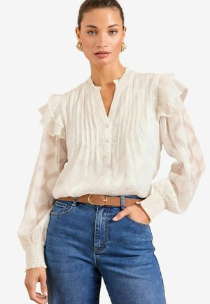 Blouse blanche avec des manches volantes, un col boutonné et un tissu texturé. Associée à un jean bleu taille haute et une ceinture marron.