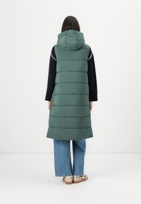 Polstret grønn ermeløs vest med hette, med horisontale sømmer, båret over en svart genser og kombinert med blå vide jeans.