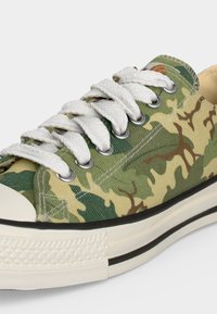 Χαμηλό sneaker με καμουφλάζ καμβά σε πράσινο και μπεζ, λευκά χοντρά κορδόνια, μεταλλικές οπές και λευκή λαστιχένια σόλα με μαύρη ρίγα.