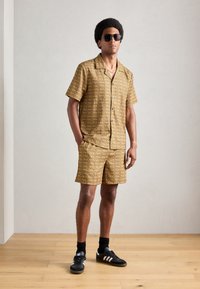 Short-sleeve shirt and shorts set in a yellow and brown repetitive pattern, with a relaxed fit. Black sneakers and sunglasses complete the look.