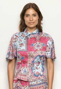 Multikleurige blouse met korte mouwen, met een patchworkontwerp van blauwe, roze en bruine bloempatronen, en een knoopsluiting aan de kraag.