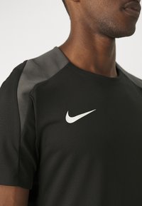 Černé sportovní tričko s šedými ramenními akcenty, vyrobené z texturované látky a s bílým logem Nike na hrudi.