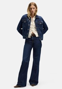 Ciemnoniebieska kurtka dżinsowa z dwoma kieszeniami na piersi, zapinana na guziki, o klasycznym kroju; zestawiona z jasnokolorową koszulą i rozszerzanymi ciemnymi jeansami.