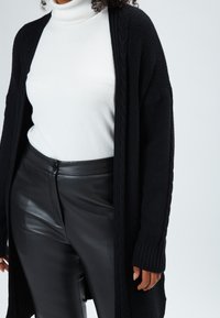 Cardigan nero a maglia con maniche lunghe sopra un dolcevita bianco, abbinato a pantaloni in pelle nera a vita alta. Texture liscia e vestibilità rilassata.