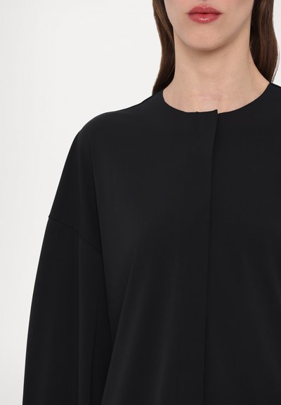 T-shirt preto com decote redondo, costuras de ombro caídas e um discreto pregueado na parte da frente. O tecido tem uma textura suave com um acabamento mate.
