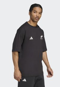 Černé bavlněné tričko s krátkým rukávem, kulatým výstřihem, bílým logem Adidas a emblémem All Blacks na hrudi, volný střih, jemná textura.