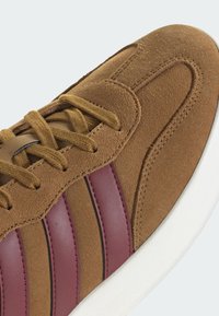 Brauner Sneaker aus Wildleder mit kastanienbraunen Lederstreifen, passenden braunen Schnürsenkeln und einer weißen Gummisohle, Nahaufnahme der Oberseite und des Zehenbereichs.