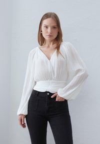 Hvid plisseret bluse med dyb V-udskæring, snøret talje og lange puffærmer, parret med højttaljede sorte denimjeans.