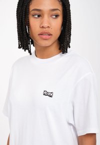 Weißes Baumwoll-T-Shirt mit kurzen Ärmeln, Rundhalsausschnitt und einem kleinen schwarzen, gestickten Logo auf der linken Brustseite.