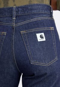 Dark blue denim jeans with yellow stitching, featuring two back pockets. A white label with a black logo is visible on the right pocket.
