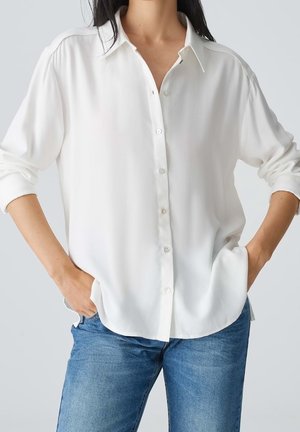 Kobieta ubrana w luźną białą koszulę na guziki z podwiniętymi rękawami i niebieskie dżinsy, ręce częściowo w kieszeniach, na jednolitym jasnym tle.