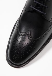 Chaussure habillée en cuir noir avec une finition lisse, un motif perforé détaillé, un design classique à bout ailé et une semelle robuste à crantage.