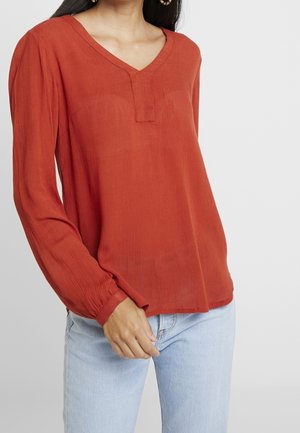 Vrouw die een roestoranje blouse met lange mouwen en een V-vormige uitsnede draagt, gecombineerd met lichtblauwe spijkerbroeken, tegen een neutrale achtergrond.
