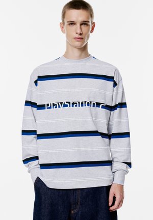 Grijze sweatshirt met lange mouwen, voorzien van horizontale strepen in blauw en zwart, met de tekst "PlayStation 2" in het wit op de voorkant.