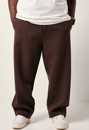 Braune weite Sweatpants aus weichem Stoff mit elastischem Bund und seitlichen Taschen, kombiniert mit weißen Low-Top-Sneakern.