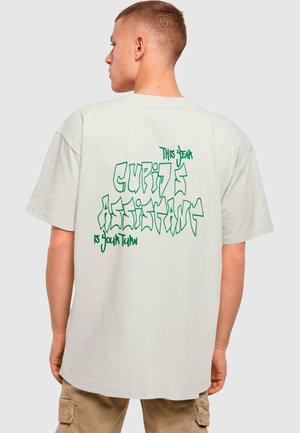 Lys grå oversized t-shirt med iøjnefaldende grøn grafisk tekst på ryggen: "Denne Kære Cupids Assistent Er Din Tur." Korte ærmer.