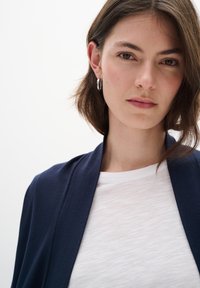 Marine strikket cardigan over en teksturert hvit T-skjorte; modellen har kort brunt hår og bruker sølvfargede hoopøreringer. Ren, nøytral bakgrunn.