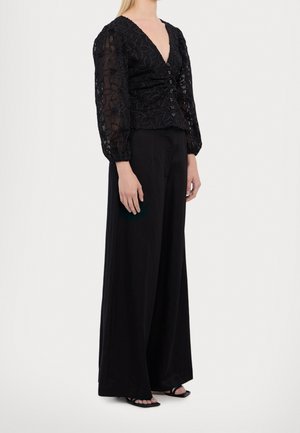 Vrouw draagt een zwarte kanten blouse met lange mouwen en V-hals met knopen, gecombineerd met zwarte, wijde broek met hoge taille en zwarte sandalen.