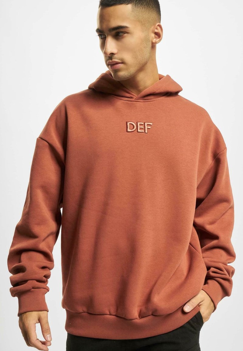 Hoodie couleur rouille en tissu doux, doté d'une poche avant, de poignets côtelés et du texte brodé "DEF" sur la poitrine.