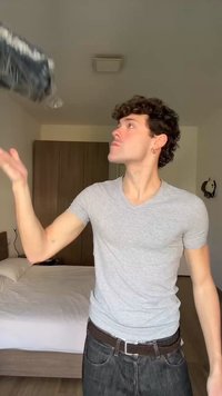 Νέος άνδρας με γκρι πουκάμισο πετάει ένα μπουκάλι νερού σε ένα υπνοδωμάτιο με κρεβάτι και ντουλάπα στο φόντο.