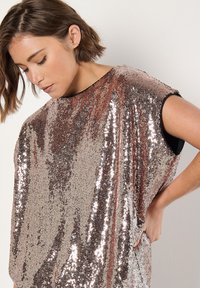 Top de lentejuelas en oro rosa con un diseño holgado y sin mangas. Presenta un escote amplio y refleja la luz con una superficie brillante y texturizada.