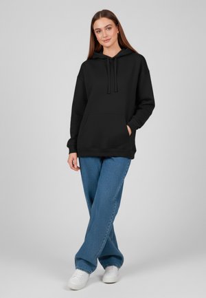 Felpa nera con cappuccio e tasca frontale, abbinata a jeans a gamba larga azzurri e sneakers bianche, su uno sfondo neutro.