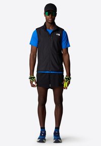 Black lightweight vest over a blue short-sleeve shirt, paired with black shorts and blue running shoes. Includes green gloves and sunglasses.