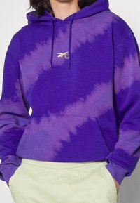 Fialová tie-dye mikina s kapucí a zlatým logem, vybavená stahovací šňůrkou a klokaní kapsou, vyrobená z měkkého fleecového materiálu.