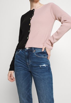 Farbblockiertes Strickpullover mit schwarzer linker Seite und pinker rechter Seite, ausgestattet mit runden grünen Knöpfen und eng anliegenden Ärmeln. Getragen mit blauen, verwaschenen Jeans.