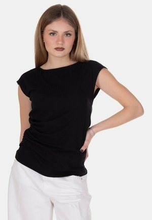 Giovane donna con lunghi capelli lisci che indossa un top nero senza maniche e pantaloni bianchi, in piedi con una mano sul fianco.