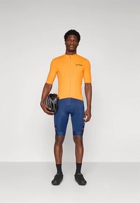 Maillot de cyclisme orange à manches courtes avec une fermeture éclair à l'avant, associé à un cuissard de cyclisme bleu. Logo texte sur les deux vêtements, casque noir tenu en main.