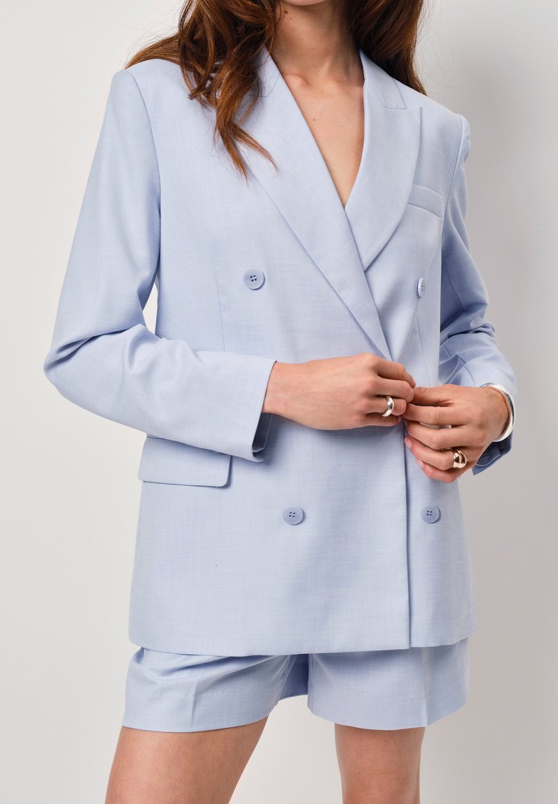 Lichtblauwe dubbelzijdige blazer met klepjes, twee voorkruiszakken en bijpassende shorts; gladde textuur en zichtbare knoopdetails.