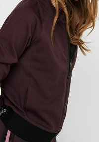 Žena v tmavě burgundské bundě na zip s černým lemem a dlouhými volně splývajícími vlasy, zachycena od ramen po pas na jednobarevném pozadí.