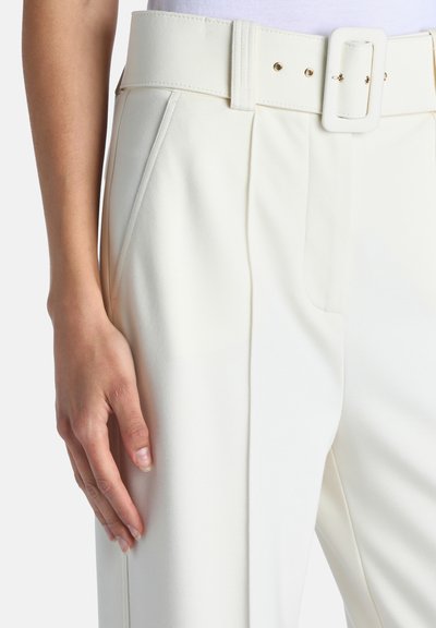 Gros plan d'une personne portant un pantalon blanc taille haute avec une large ceinture et une main reposant à côté de la cuisse.