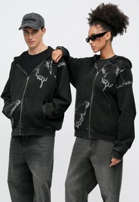 Μαύρο φούτερ με φερμουάρ από μαλακό ύφασμα, με γκρι γραφικά πεταλούδας στους ώμους και τις μπροστινές τσέπες, σε συνδυασμό με σκούρο γκρι τζιν.