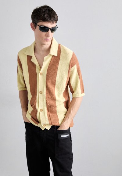 Kurzarm-Hemd mit Knöpfen in Gelb- und Braunstreifen, strukturierter Stoff, mit zwei frontalen Taschen, kombiniert mit schwarzen Hosen und Sonnenbrille.