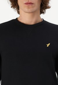 Zwarte sweatshirt van zacht materiaal, met een ronde halslijn en een klein gouden vogellogo op de linkerkant van de borst.