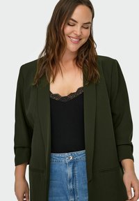 Smaidīga sieviete ar vaļīgiem brūniem matiem, tērpusies tumši zaļā žaketē, melnā topā ar mežģīņu apdari un augsta vidukļa zilās džinsa biksēs pret baltu fonu.