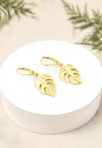 Örhängen i guldbladform med utskärningar och lyftstängning, presenterade på en rund vit displaypedestal.