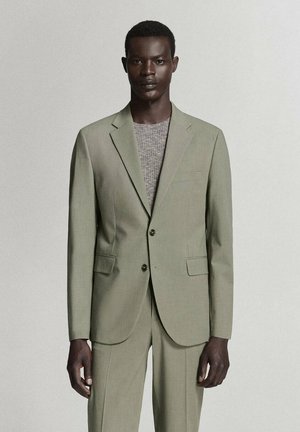 Hombre vistiendo un traje verde claro hecho a medida con dos botones y un suéter gris de lana debajo, de pie frente a un fondo claro y simple.