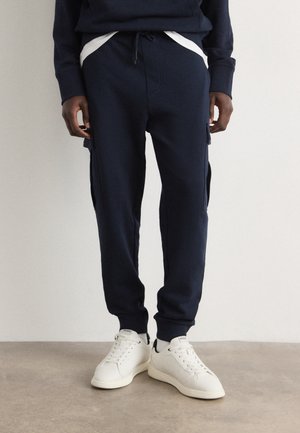 Person trägt marineblaue Cargo-Jogginghosen mit weißem Kordelzug und weißen Sneakers und steht auf einem hellen Boden vor einer schlichten weißen Wand.