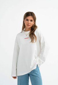 Weißes langärmeliges Sweatshirt mit Rundhalsausschnitt und rotem "MIRONS"-Text auf der Brust. Kombiniert mit hellblauen Jeans.